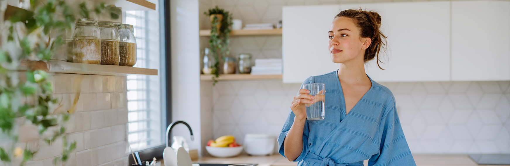 Frau trinkt Wasser in der Küche Frau in blauem Bademantel steht in moderner Küche und hält ein Glas Wasser, neben Spüle mit Wasserhahn